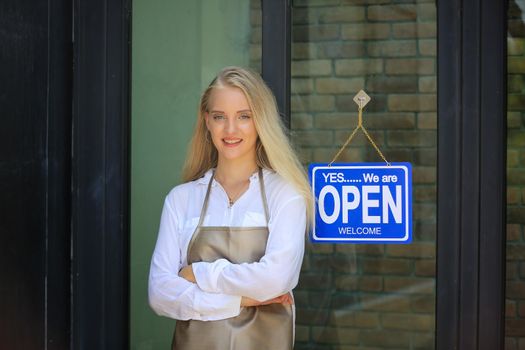 Beautiful blonde hair girl standing by open shop sign plate at door with confidence in front of coffee shop.