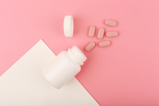 Creative flat lay with nutritional supplement or vitamins for woman. Vitamins for nail and hair growth or supplement for beautiful skin. Opened medication bottle with spilled pink pills on pink background. 