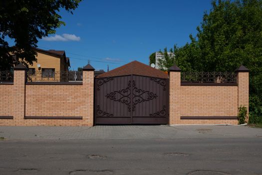 Light brick fence with iron wrought iron gates on the street against a background of wood and blue sky.