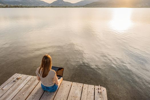 Lonely girl expressing her creativeness writing romance tales book on a wooden pier or jetty at sunset or dawn. New job opportunity at modern times using laptop and wifi internet connection technology