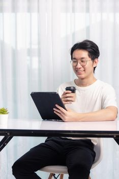 Happy Asian young business handsome man work from home office wear glasses, t-shirt comfortable he smiling and using a black modern smart digital tablet computer to read email or learning on desk