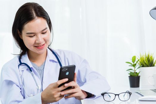 Asian doctor young beautiful woman smiling using working or holding with smart mobile phone and laptop computer at hospital desk office, technology healthcare medical concept