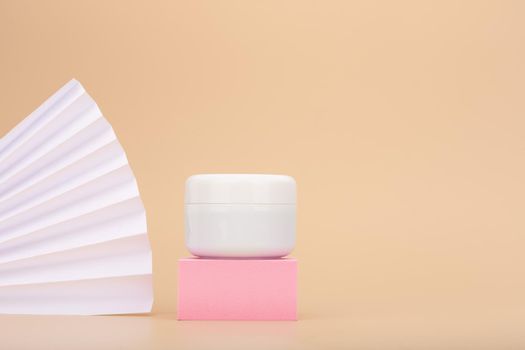 Cosmetic cream jar on pink podium with white waver next to it against beige background with copy space. Face cream, body scrub or hair mask on pastel beige background. Concept of beauty products 