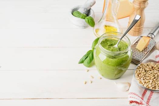 Italian cuisine. Preparing homemade italian pesto sauce. Homemade pesto sause in a glass jar with ingredients on white wooden table, copy space