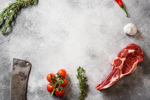 Raw black angus tomahawk steak or Spencer steak cut with the meat american cleaver knife over grey stone table slate cutting top view frame with seasonings, tomatoes, garlic, rosemary. Space for text.
