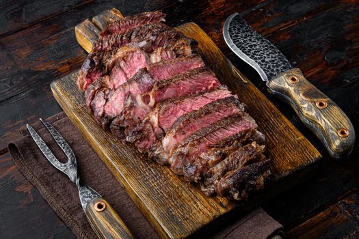 Juicy medium beef Rib Eye steak slices set, on wooden serving board, with meat knife and fork, on old dark wooden table background