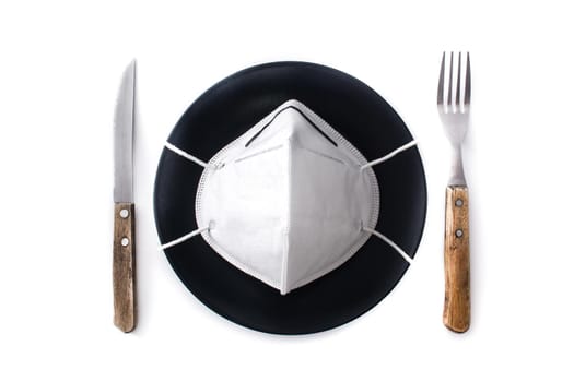 Protective face mask on a black plate,fork and knife. Eating at a restaurant during a coronavirus pandemic concept.