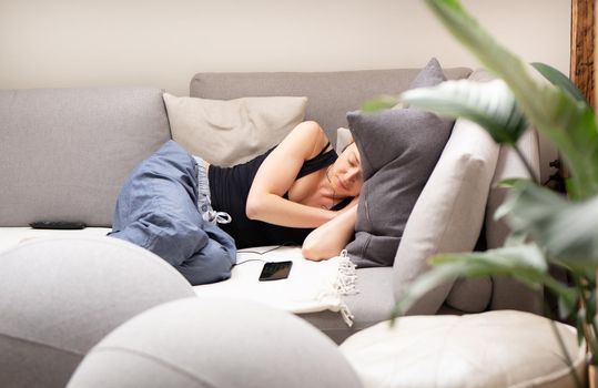 Tired young woman taking a nap at home lying on a comfortable sofa with big cushions in living room with a mobile phone lying next to her. Healthy lifestyle concept.