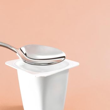 Yogurt cup and silver spoon on beige background, white plastic container with yoghurt cream, fresh dairy product for healthy diet and nutrition balance.