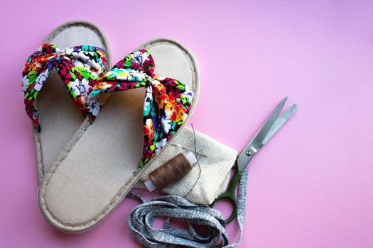 Thread needle scissors and colored home women's slippers on a pastel paper-pink background. Top view. Copy space.