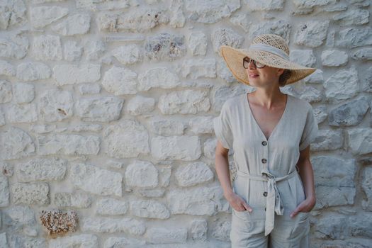 Portrait of beautiful cheerful blonde woman wearing one piece sundress and summer hat, standing in front of old medieval stone wall. Summer vacation portrait concept. Copy space.