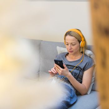 Young thoughtful woman sitting indoors at home living room sofa using social media on phone for video chatting and staying connected with her loved ones. Stay at home, social distancing lifestyle.