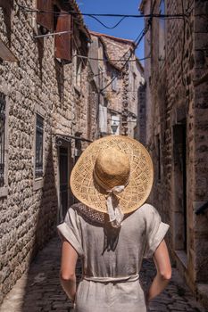 Beautiful blonde young female traveler wearing straw sun hat sightseeing and enjoying summer vacation in an old traditional costal town at Adriatic cost, Croatia. Rear view against the old stone town.