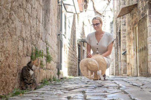 Beautiful blonde young female traveler wearing straw sun hat caressing cat while enjoying summer vacation in an old traditional costal town at Adriatic cost, Croatia.