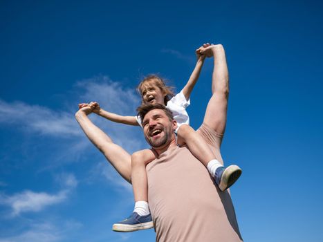 Little girl sit on the neck of father with raised arms over blue sky background