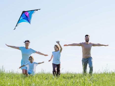 Happy young parents and children in casual clothes running with outstretched arms while enjoying time together on green meadow flying a kite