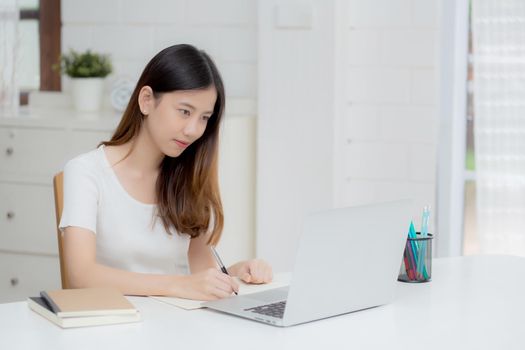 Young asian woman writing on notebook for planning working and using laptop computer on desk at home, girl notes about finance, female study and learning, business and communication concept.