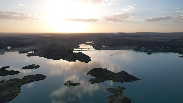 A speedboat sails along the strait at sunset. Top view from the drone. A long plume on the water. You can see the bridge and the rocky shore. The water reflects clouds and the sky. Kapchagai reservoir