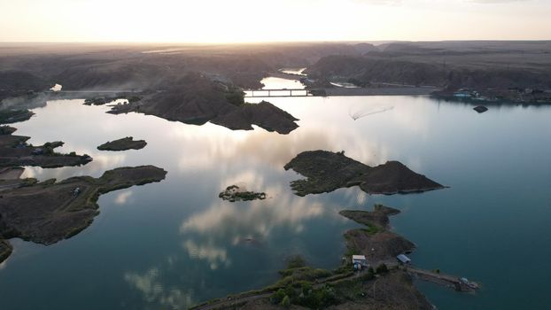 A speedboat sails along the strait at sunset. Top view from the drone. A long plume on the water. You can see the bridge and the rocky shore. The water reflects clouds and the sky. Kapchagai reservoir