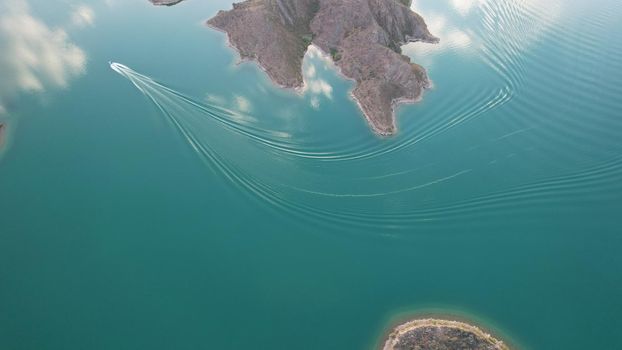 A speedboat sails along the strait at sunset. Top view from the drone. A long plume on the water. You can see the bridge and the rocky shore. The water reflects clouds and the sky. Kapchagai reservoir