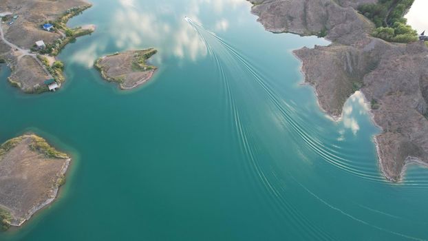 A speedboat sails along the strait at sunset. Top view from the drone. A long plume on the water. You can see the bridge and the rocky shore. The water reflects clouds and the sky. Kapchagai reservoir