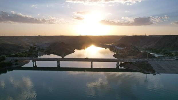 Sunset with a view of the causeway and the bridge over the strait. Cars drive on the bridge. The water reflects the sun, sky and clouds. There are houses on the rocky shore. Kapchagai reservoir.