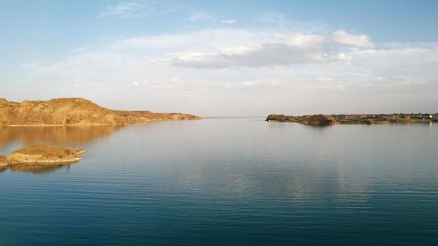 Flying over a lake with islands and rocks. Green water reflects the rays of the sun. Trees and bushes grow on the rocky ground, and houses are built. White clouds. Kapchagai Reservoir, Kazakhstan.