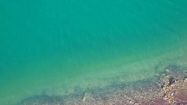 Top view of green water and small waves. The background of the water shimmers in the rays of the sun with highlights. White foam is visible. The gradient of the lake and the beach. Rocks on the bottom