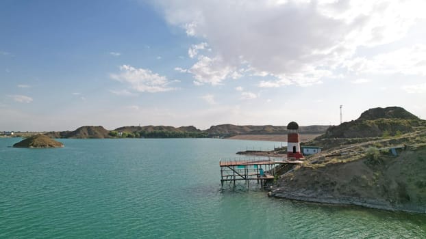 White and red lighthouse on the shore of the lake. The green water of the reservoir reflects the sky and clouds. Rocky beach. In some places, trees and bushes grow. Shtul. Kapchagai, Kazakhstan.