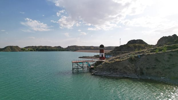 White and red lighthouse on the shore of the lake. The green water of the reservoir reflects the sky and clouds. Rocky beach. In some places, trees and bushes grow. Shtul. Kapchagai, Kazakhstan.