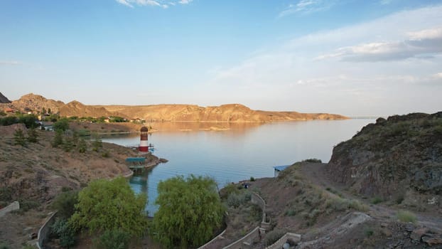 White and red lighthouse on the shore of the lake. The green water of the reservoir reflects the sky and clouds. Rocky beach. In some places, trees and bushes grow. Shtul. Kapchagai, Kazakhstan.