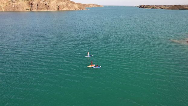 A group of people ride sup surfing in the lake. Aerial view from the drone of the green water and rocky beaches. Inflatable sapboard board. Trees and bushes grow on the bank of Kapchagai.