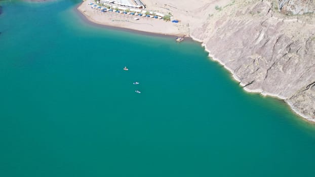 A group of people ride sup surfing in the lake. Aerial view from the drone of the green water and rocky beaches. Inflatable sapboard board. Trees and bushes grow on the bank of Kapchagai.