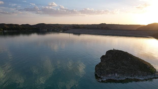 A small island in the middle of the bay. Mirror water reflects the sun, clouds, and sky. Birds fly. In the distance, you can see the road, cars, trees and houses. Kapchagai Reservoir, Kazakhstan.