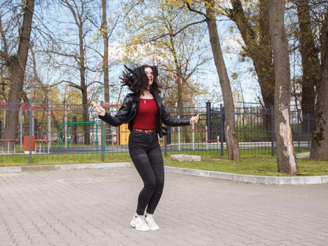 young brunette girl in black jacket and jeans jumping rope outdoor on sunny spring day