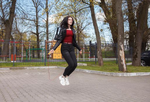young brunette girl in black jacket and jeans jumping rope outdoor on sunny spring day