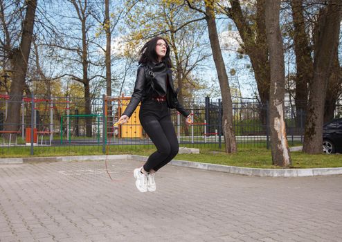 young brunette girl in black jacket and jeans jumping rope outdoor on sunny spring day