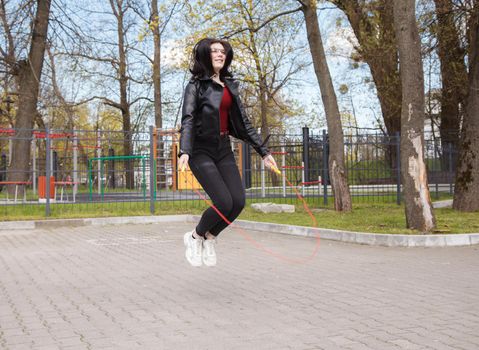 young brunette girl in black jacket and jeans jumping rope outdoor on sunny spring day