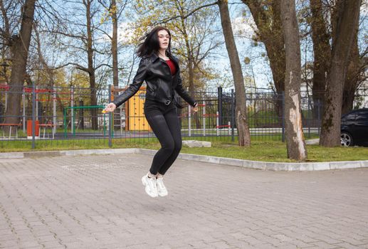 young brunette girl in black jacket and jeans jumping rope outdoor on sunny spring day