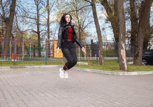 young brunette girl in black jacket and jeans jumping rope outdoor on sunny spring day