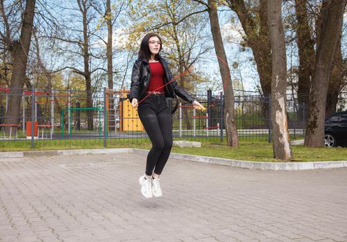 young brunette girl in black jacket and jeans jumping rope outdoor on sunny spring day