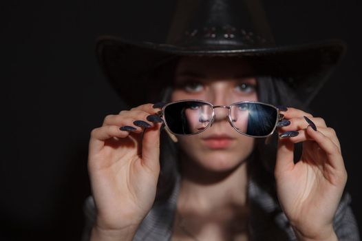 portrait of a young beautiful brunette girl in jacket and hat with glasses in studio on black background