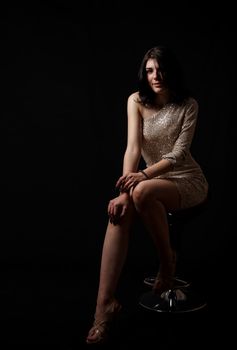 young beautiful brunette girl in bright dress posing sitting on chair in the studio on black background