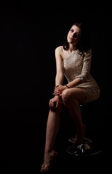 young beautiful brunette girl in bright dress posing sitting on chair in the studio on black background