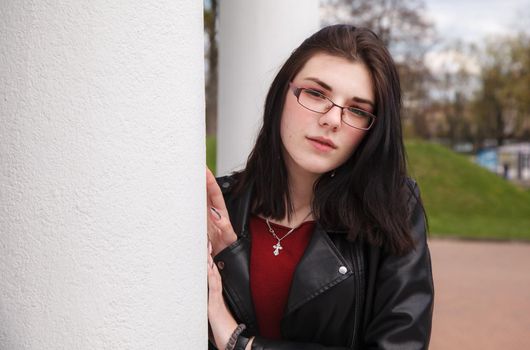 young brunette beautiful in black leather jacket girl peeking out from behind pillar in city park. outdoor sunny spring day