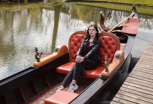 young brunette girl in black jacket and jeans sits in boat on lake in city park on sunny day