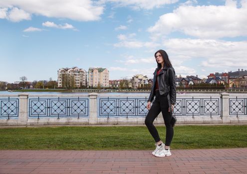 young beautiful brunette girl in black jacket and jeans stands on the embankment near lake in city park on sunny spring day