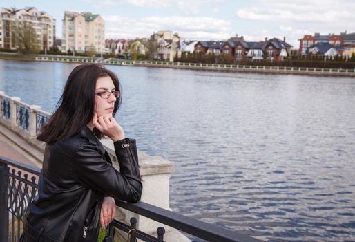 young beautiful brunette girl in black jacket and jeans stands on the embankment near lake in city park on sunny spring day