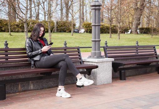 young girl in black jacket and jeans looking at smartphone display while sitting on bench in city park on sunny spring day