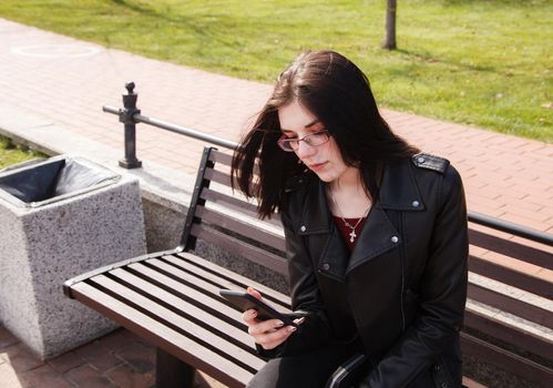 young girl in black jacket and jeans looking at smartphone display while sitting on bench in city park on sunny spring day. outdoor closeup